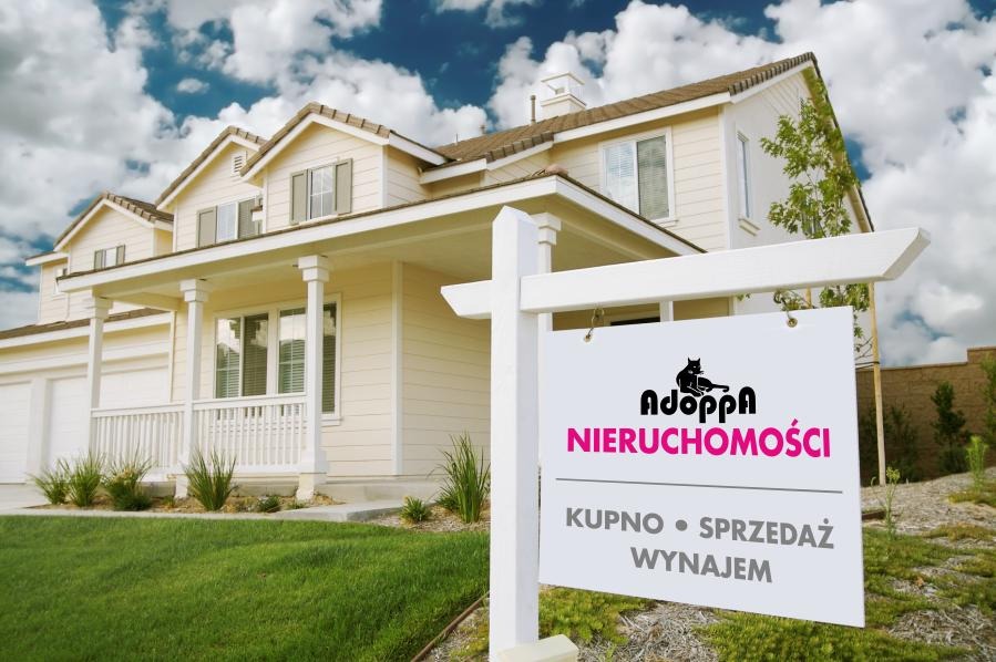 Dom jednorodzinny z beżową elewacją i białymi oknami, na pierwszym planie tablica agencji nieruchomości z logo czarnego kota i napisem 'Kupno • Sprzedaż • Wynajem'.