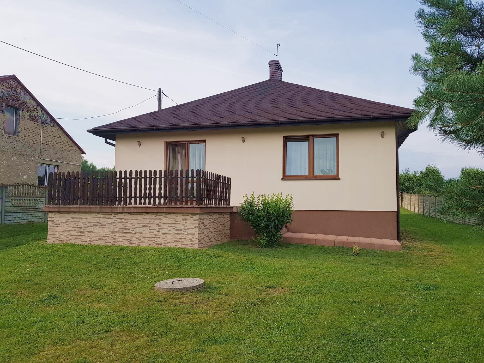 Parterowy dom jednorodzinny z brązowym dachem i drewnianym ogrodzeniem tarasu, widok z ogrodu w letni dzień.