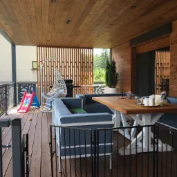 Zadaszony taras z drewnianym sufitem i ścianami, meble ogrodowe z szarej tapicerki, drewniany stół na białej podstawie, wiszące krzesło, widok na ogród, pies siedzący na ławce przy stole, elementy...