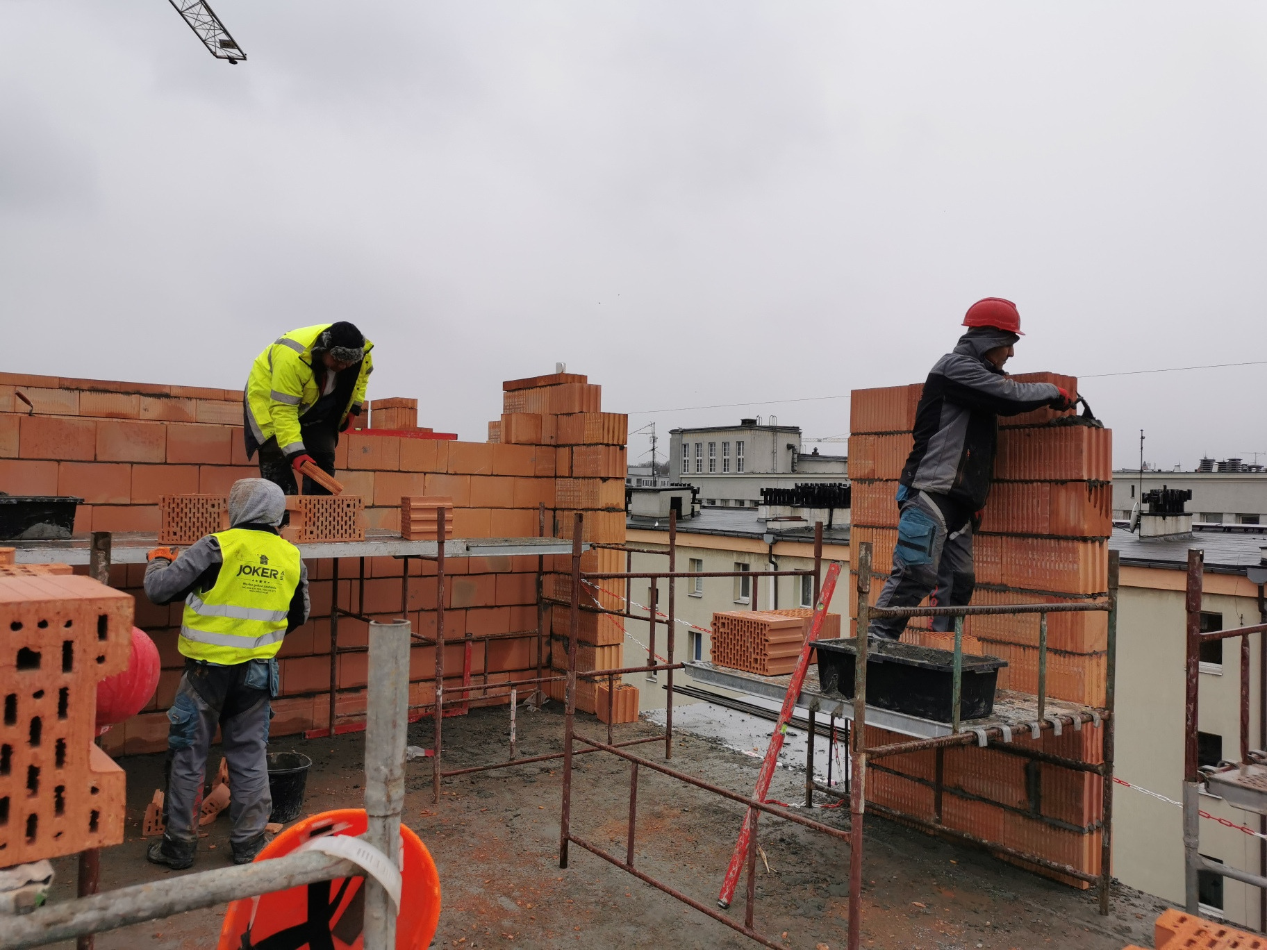 Murarze w trakcie wznoszenia ścian z czerwonej cegły na rusztowaniu, widoczne budynki w tle
