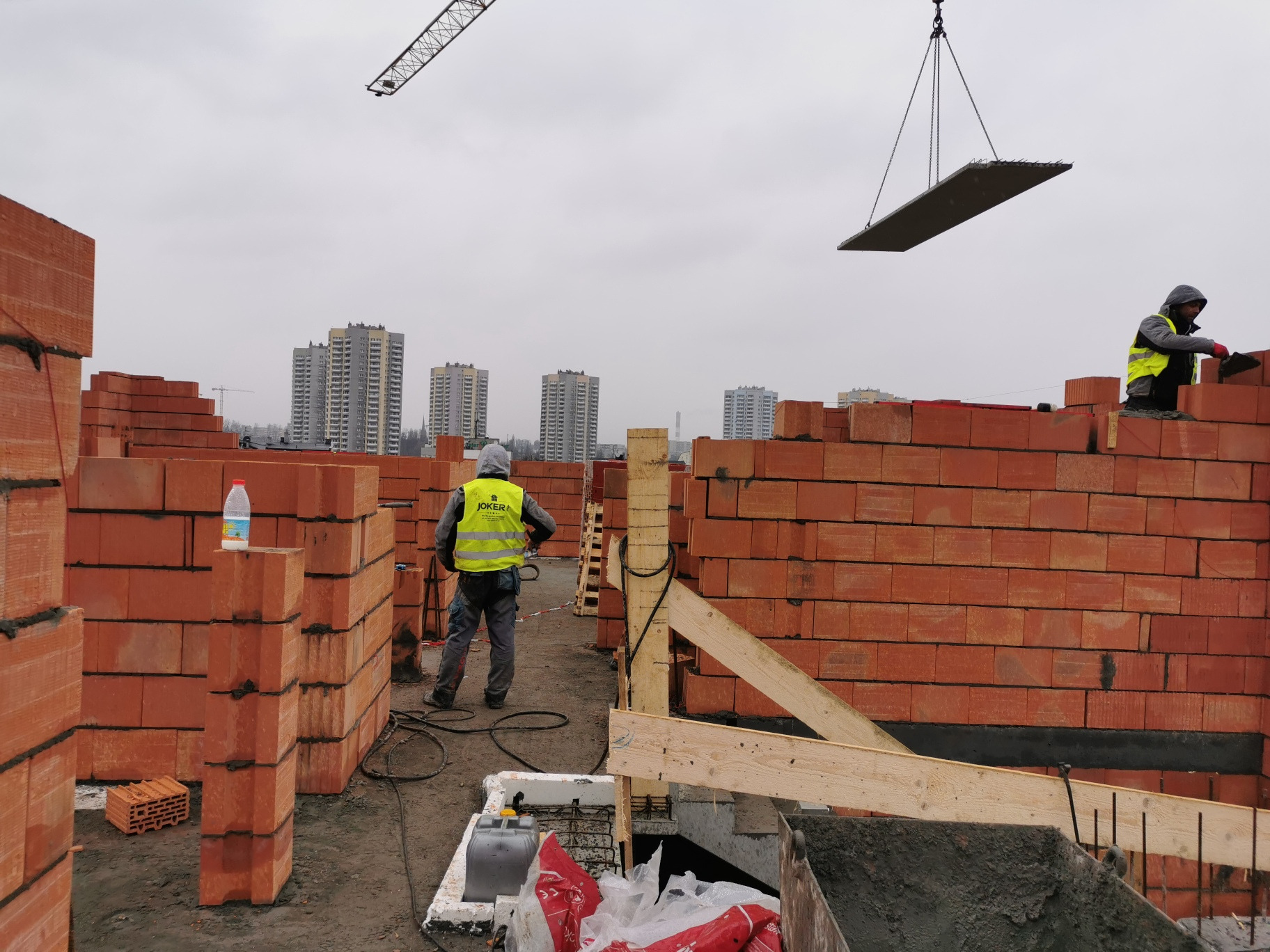 Budowa domu murowanego: robotnicy w kamizelkach odblaskowych układają cegły, dźwig podnosi betonowy element stropu, w tle widoczne budynki mieszkalne i pochmurne niebo.