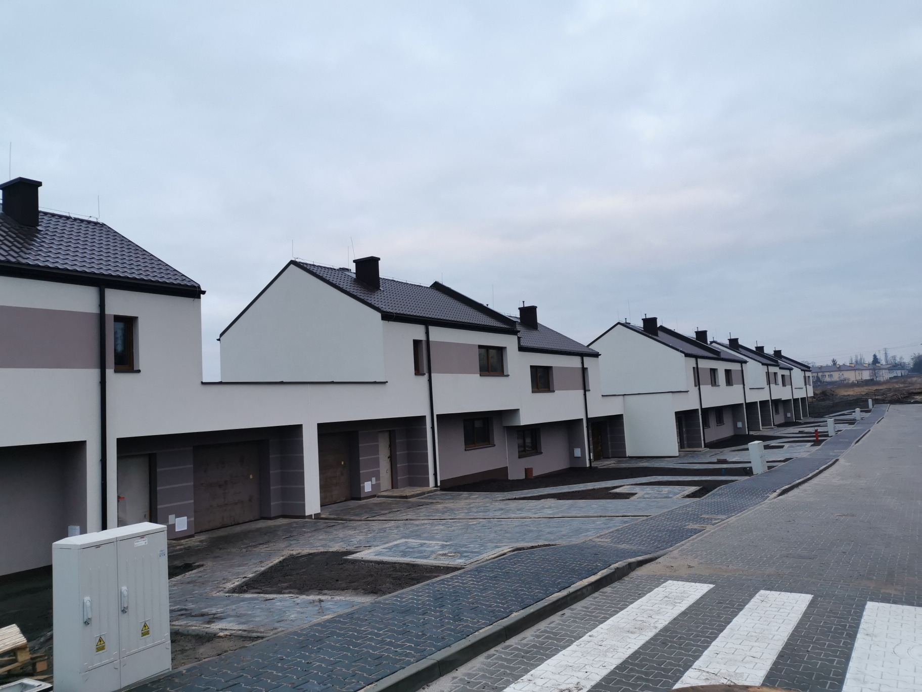 Szereg nowo wybudowanych, dwukondygnacyjnych domów z garażami, wykończonych w bieli i szarości, z ciemnymi dachami i oknami, na tle pochmurnego nieba i przygotowanego terenu.