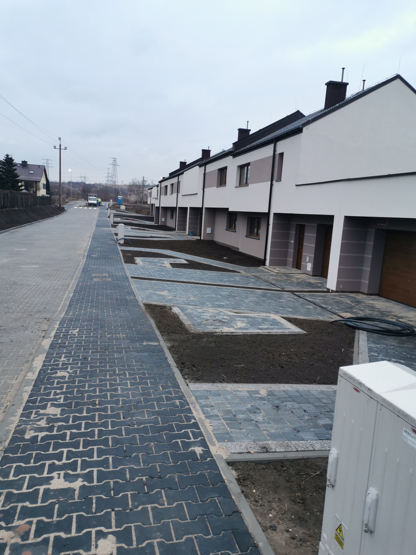 Rząd nowych, dwukondygnacyjnych domów szeregowych z płaskimi dachami i garażami, widok z perspektywy chodnika z ciemnej kostki brukowej, teren wokół domów przygotowany pod trawniki.