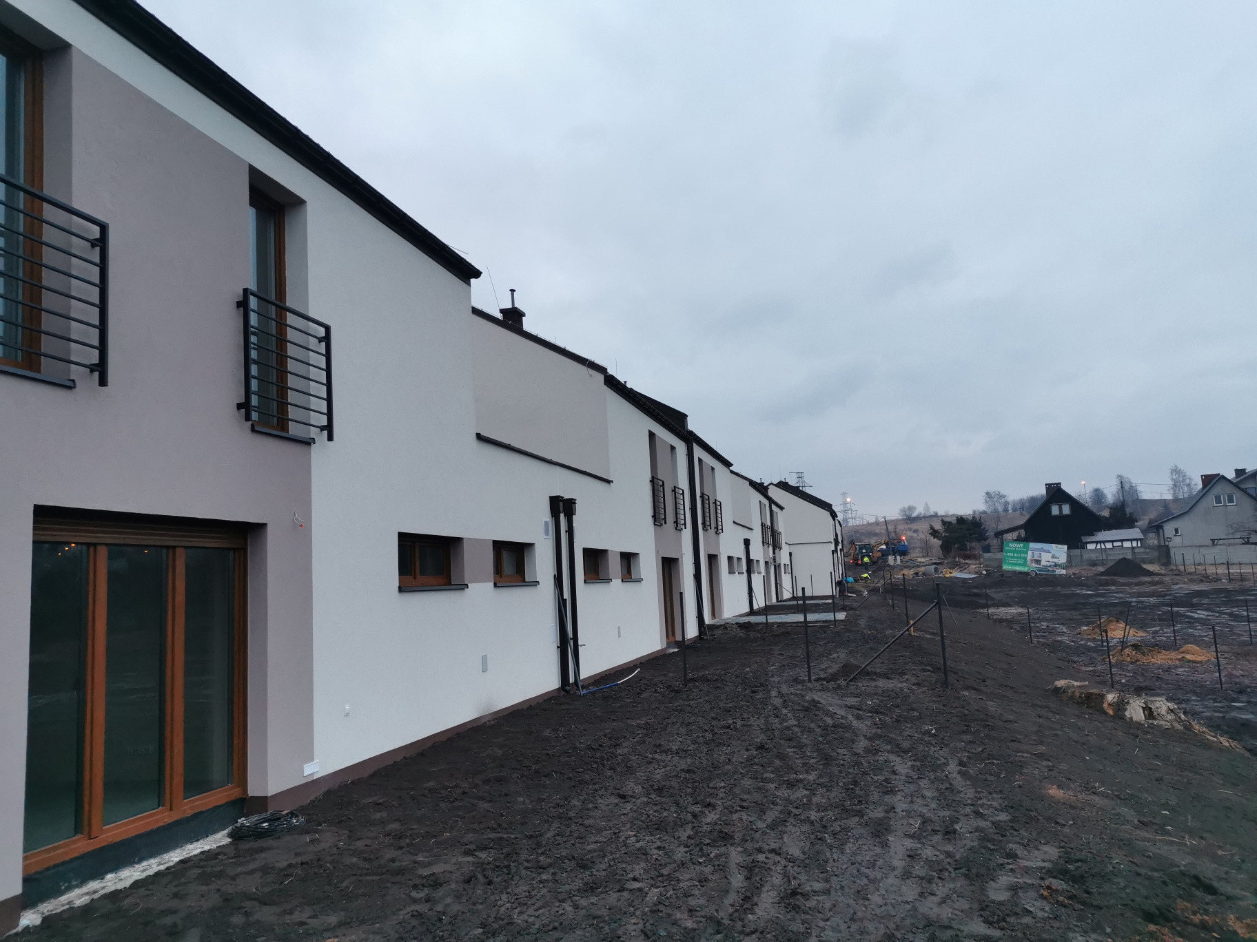 Rząd nowo wybudowanych domów jednorodzinnych z balkonami i ciemną stolarką okienną, widoczny teren wokół budynków w trakcie zagospodarowywania, błotniste podłoże.