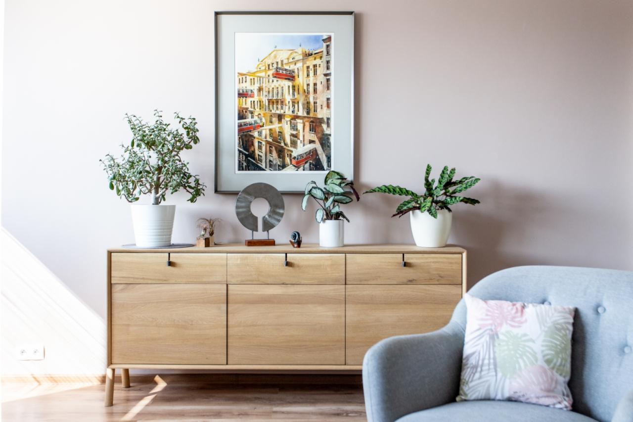 Jasny salon z drewnianą komodą, na której stoją rośliny doniczkowe, metalowa dekoracja i obraz z miejskim pejzażem z tramwajami; w tle ściana w odcieniu pudrowego różu i fragment szarego fotela.