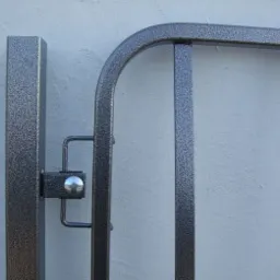 Metalowy panel ogrodzeniowy z widocznym mocowaniem i fakturą w kolorze szaro-srebrnym na tle jasnej ściany.