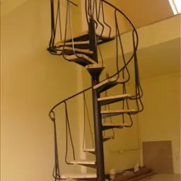 Schody spiralne z czarną, kutą balustradą i jasnymi, drewnianymi stopniami, widoczne z dołu, w jasnym pomieszczeniu z beżowymi ścianami i skośnym sufitem z oknem dachowym.