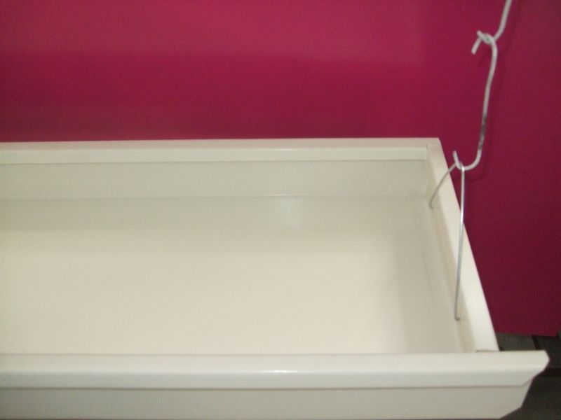 Wysuwana szuflada w kolorze złamanej bieli, przymocowana do regału za pomocą metalowych zawieszek, na tle ściany w kolorze fuksji.