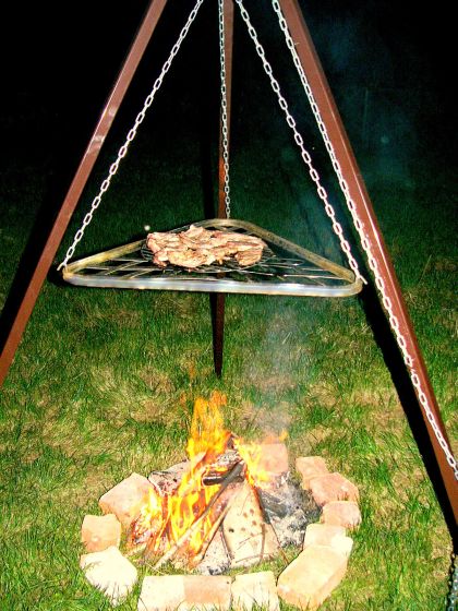 Ognisko otoczone cegłami na trawie, nad nim trójnóg z rusztem i mięsem na grillu, oświetlone płomieniami ognia.