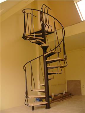 Schody spiralne z czarną, kutą balustradą i jasnymi, drewnianymi stopniami, widoczne z dołu, w jasnym pomieszczeniu z beżowymi ścianami i skośnym sufitem z oknem dachowym.