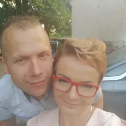 Selfie murarza i kobiety w czerwonych okularach na tle świeżo otynkowanego budynku i samochodu.