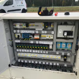 Otwarta szafa sterownicza z widocznymi elementami instalacji elektrycznej, zabezpieczeniami i przewodami, z torbą narzędziową na górze, obok samochód firmy elektrycznej w Gryfinie.