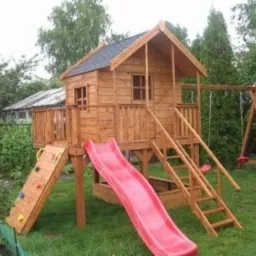 Drewniany plac zabaw z domkiem na palach, czerwoną zjeżdżalnią, ścianką wspinaczkową i huśtawką na tle zielonego ogrodu.