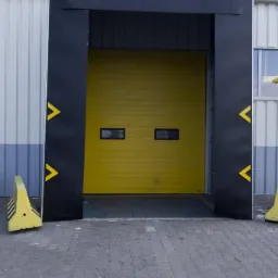 Żółta brama przemysłowa z dwoma małymi okienkami, otoczona czarną ramą z żółtymi strzałkami, widoczna z zewnątrz budynku przemysłowego.
