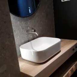 Nowoczesna łazienka z białą, prostokątną umywalką nablatową, chromowaną baterią, okrągłym lustrem i drewnianą szafką z dekoracyjnymi elementami.