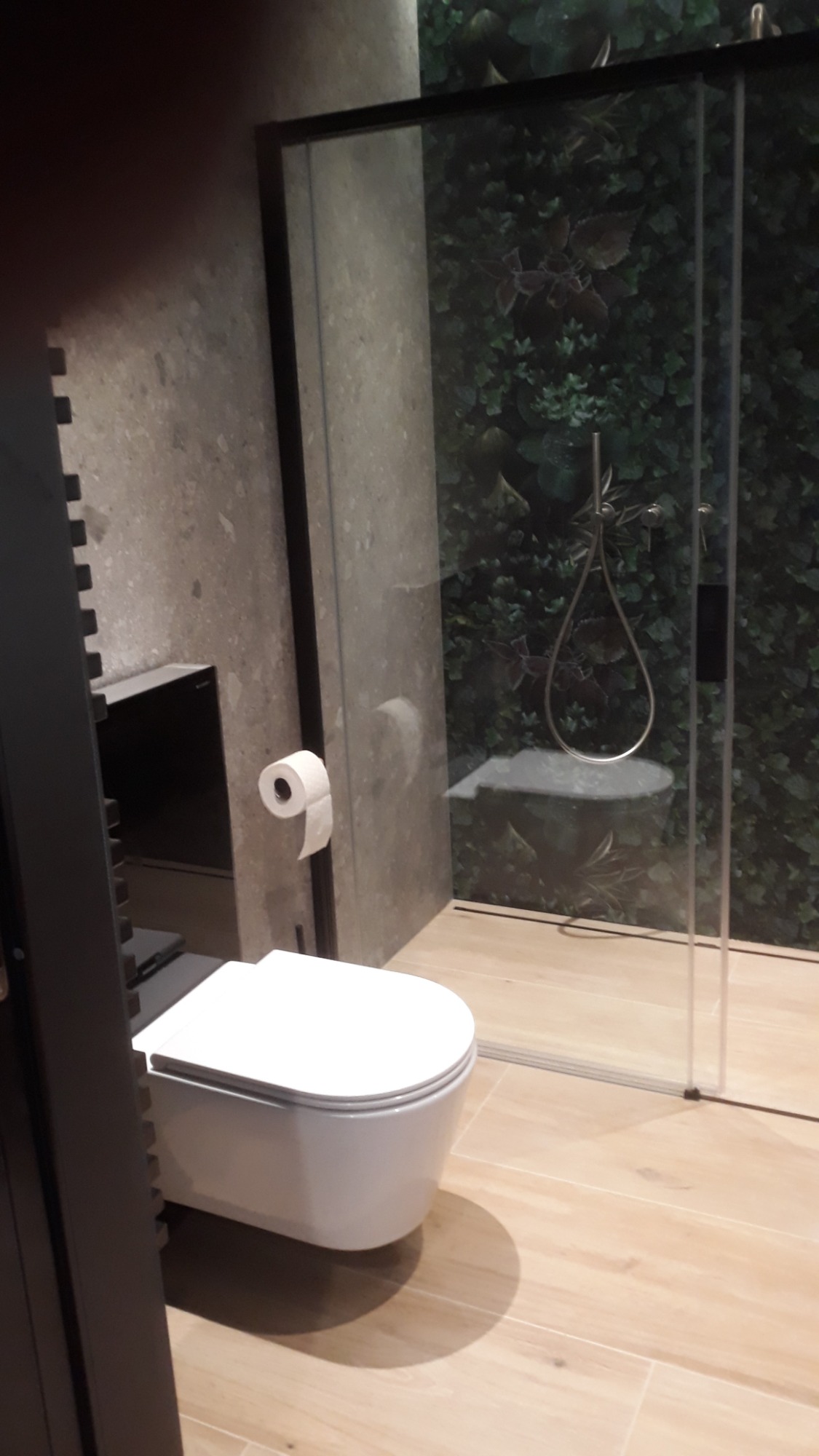 Nowoczesna toaleta podwieszana w łazience z kabiną prysznicową typu walk-in, wykończoną płytkami imitującymi drewno i ścianą z motywem roślinnym.