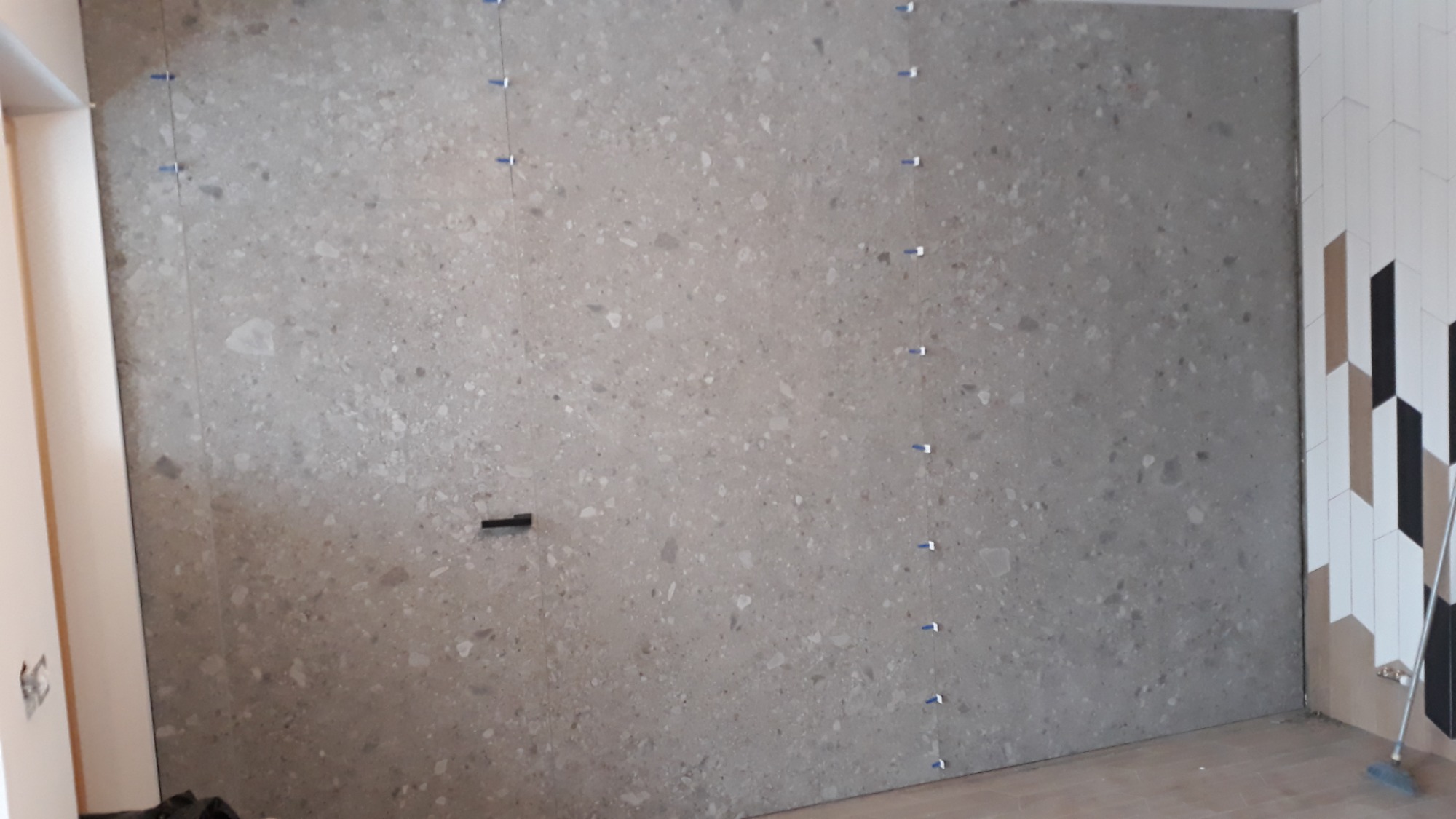 Ściana z szarych płytek imitujących kamień, z systemem poziomowania płytek i fragmentem ściany z dekoracyjnymi płytkami w romby w odcieniach bieli, beżu i czerni.