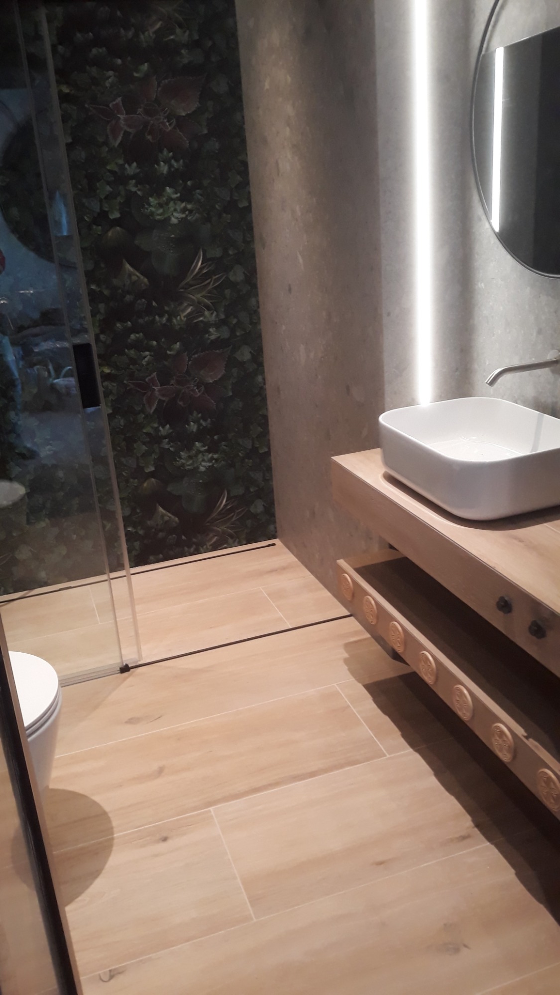 Nowoczesna łazienka z drewnianą szafką pod umywalką, dekoracyjnymi elementami na półce i ścianą z motywem roślinnym za kabiną prysznicową.