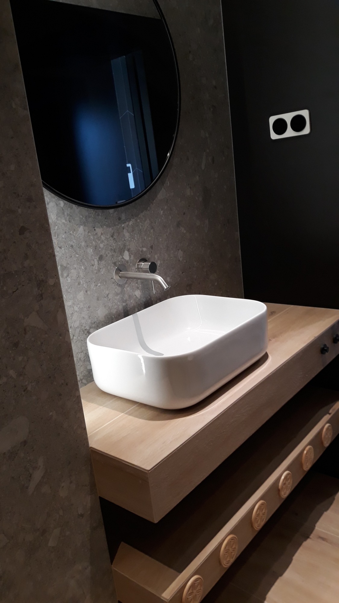 Nowoczesna łazienka z białą, prostokątną umywalką nablatową, chromowaną baterią, okrągłym lustrem i drewnianą szafką z dekoracyjnymi elementami.