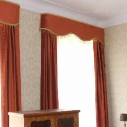 Rustykalne, pomarańczowe zasłony z lambrekinem zdobionym złotymi frędzlami w eleganckim wnętrzu z wzorzystą tapetą i drewnianą komodą.