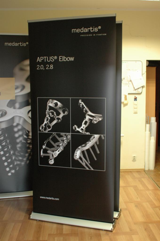 Czarny rollup reklamowy Medartis APTUS Elbow 2.0, 2.8 z ilustracjami implantów ortopedycznych na drewnianej podłodze, widoczny fragment drugiego rollupu w tle.