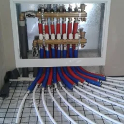 Rozdzielacz ogrzewania podłogowego w metalowej szafce, rury czerwone i niebieskie prowadzone na matę z rastrem, widoczne obejmy mocujące rury.