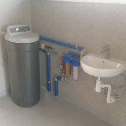 Szary zmiękczacz wody z filtrami wstępnymi i umywalką zamontowany w pomieszczeniu technicznym z szarą podłogą i ścianami w kropki.
