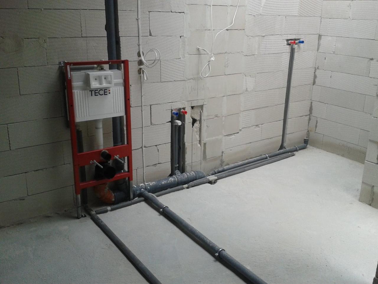 Surowa łazienka w trakcie instalacji: stelaż podtynkowy TECE z czerwoną ramą, szare rury kanalizacyjne prowadzone po podłodze i ścianach z betonu komórkowego, widoczne przyłącza wody z zaworami.