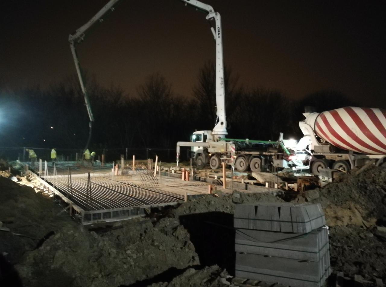 Nocna budowa fundamentów pod budynek: widoczny szalunek, zbrojenie, beton pompowany przez pompę na samochodzie oraz betoniarka na tle ciemnego nieba i drzew.