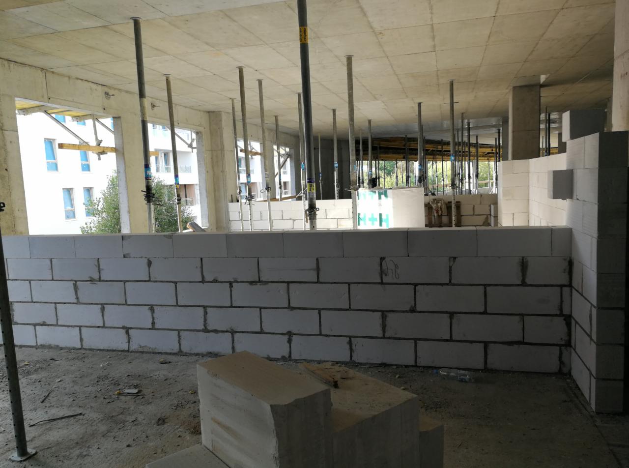 Wznoszenie ścian z białych bloczków betonu komórkowego w surowym stanie, widoczne podpory stropu i budynki w tle.