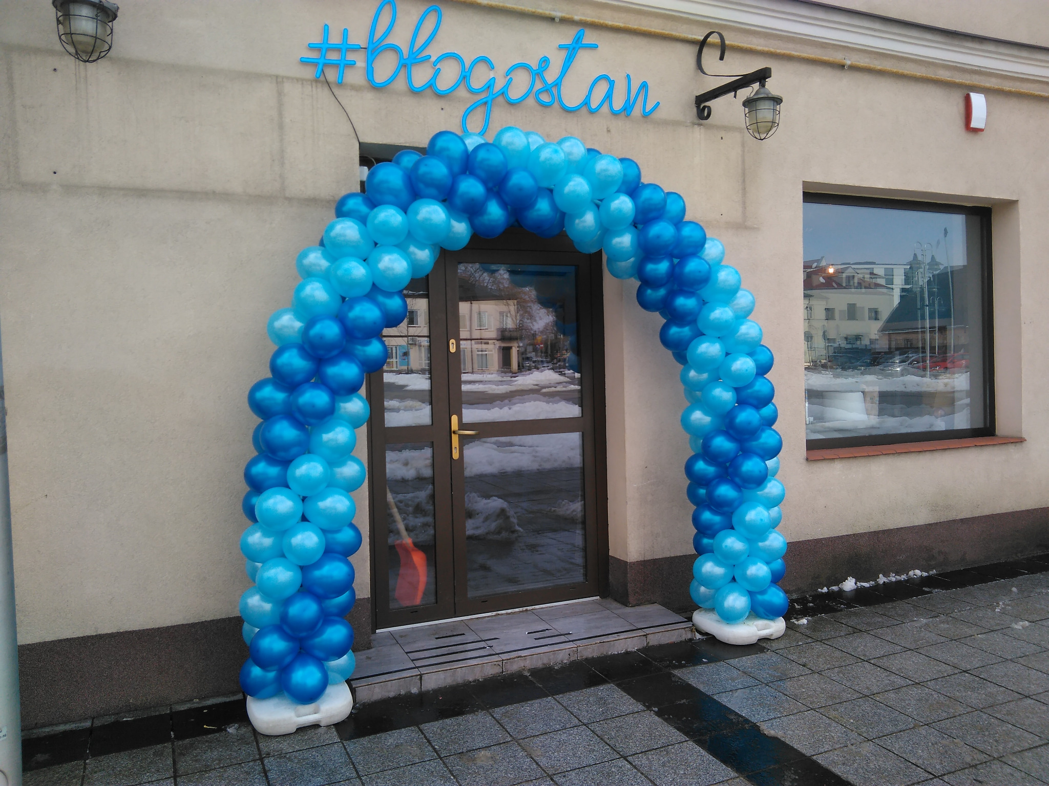 Wejście do budynku udekorowane łukiem z niebieskich i błękitnych balonów, nad wejściem neonowy napis #blogostan.