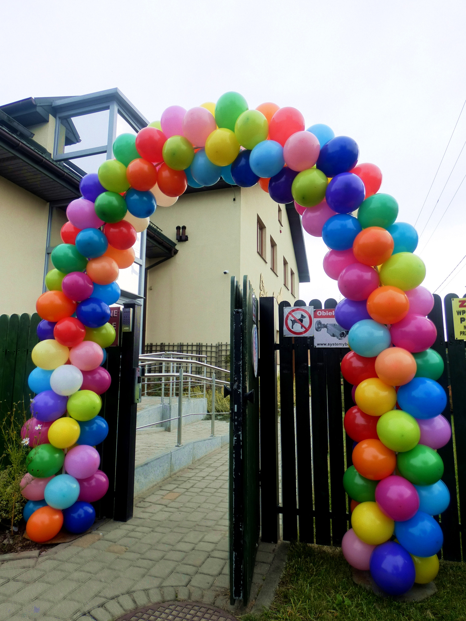 Kolorowy łuk z balonów przy wejściu na posesję z zielonym płotem i budynkiem w tle.