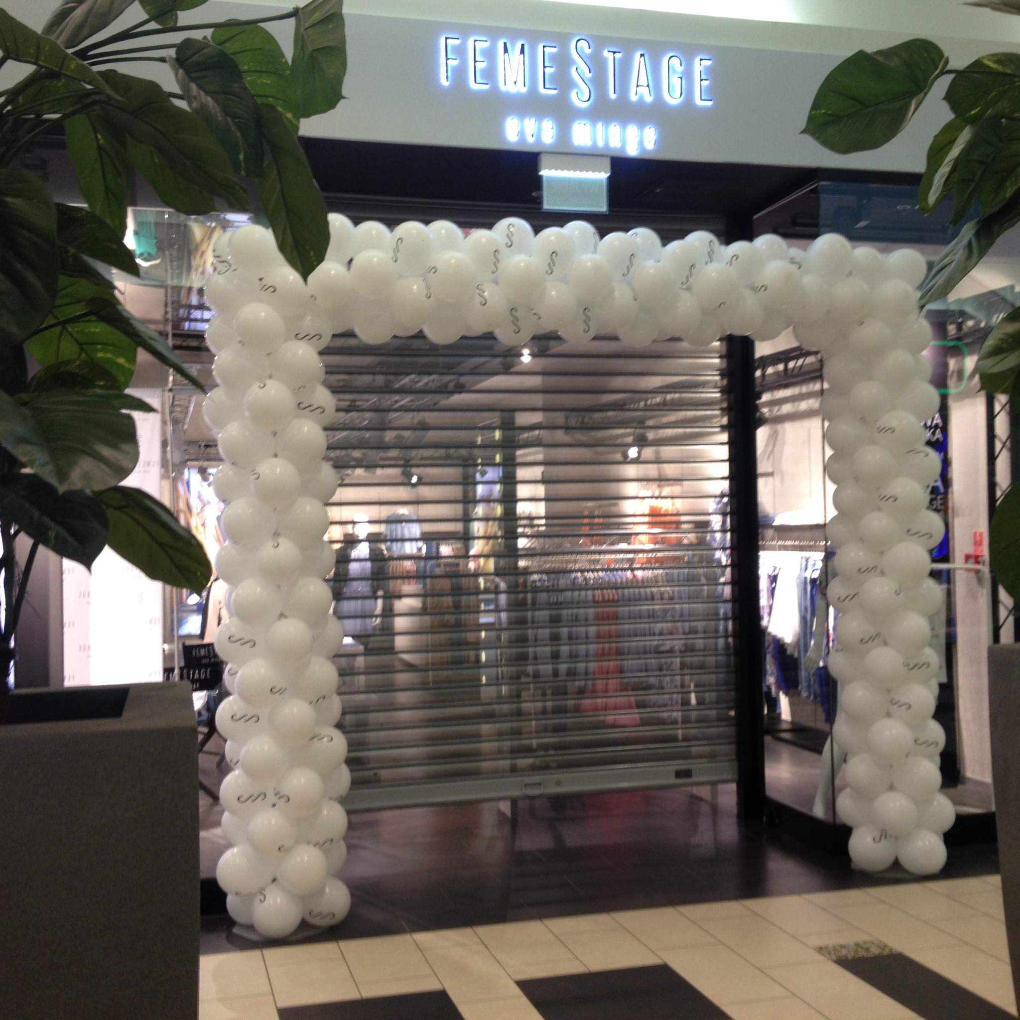 Biały łuk balonowy z literą 'S' przed zamkniętym sklepem Femestage w centrum handlowym, widoczne rośliny doniczkowe.