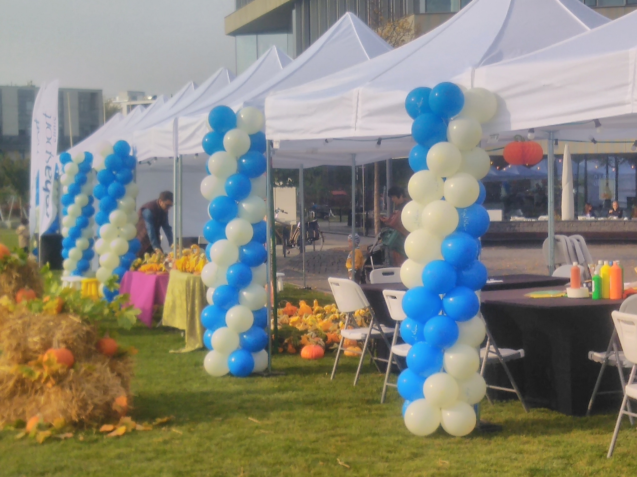 Dekoracja plenerowa z białych namiotów, kolumn z niebiesko-białych balonów, dyń i słomy, przygotowana na imprezę.