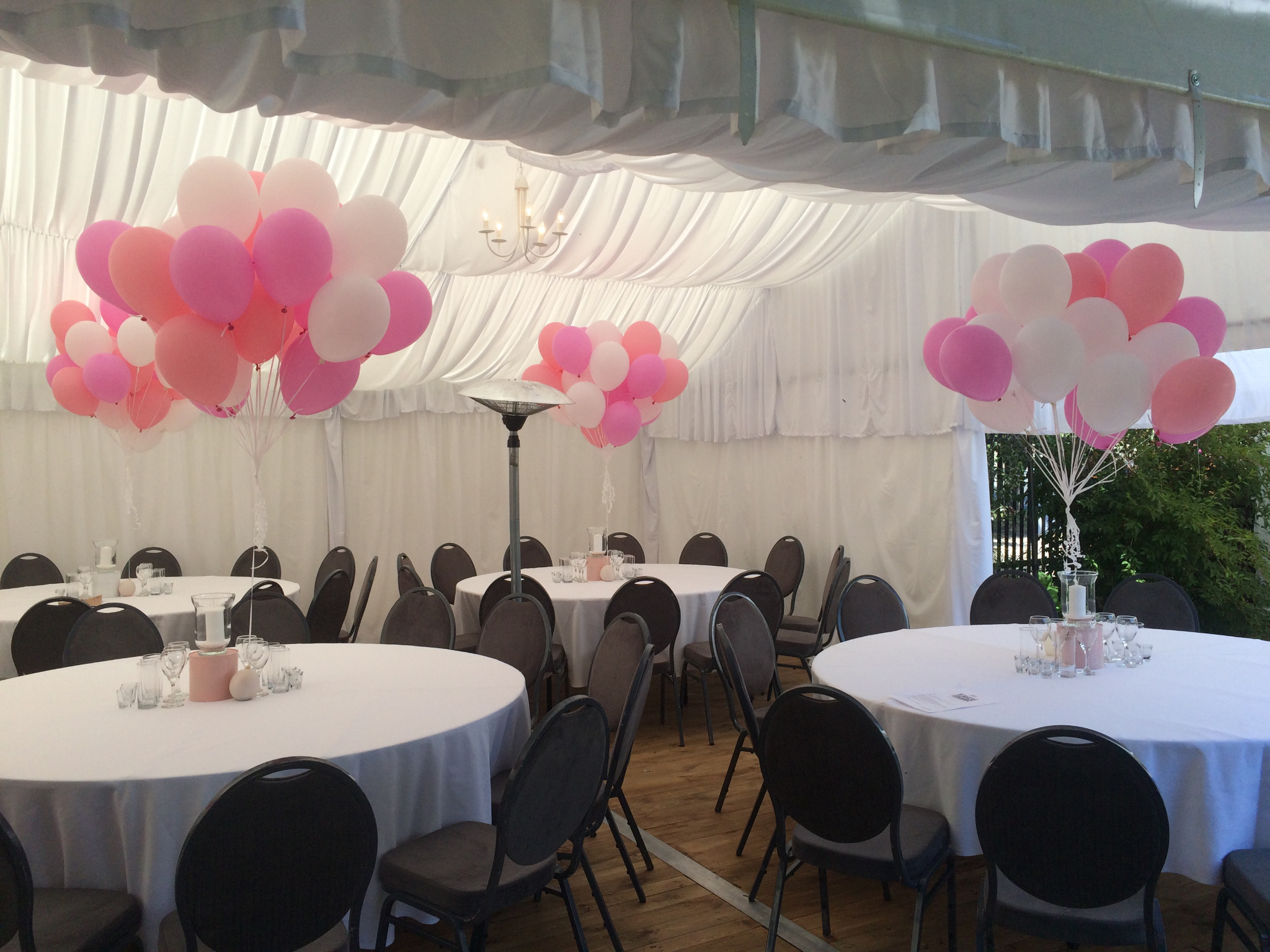 Uroczyste wnętrze namiotu z okrągłymi stołami, ciemnymi krzesłami i dekoracjami z różowych i białych balonów pod białym, drapowanym sufitem.