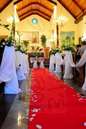 Czerwony dywan z białymi płatkami róż w kościele, dekoracje kwiatowe przy ławkach, przygotowania do ceremonii ślubnej.