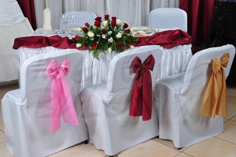 Uroczysty stół z białym obrusem i kompozycją z czerwonych i białych róż, krzesła z białymi pokrowcami ozdobione różowymi, bordowymi i złotymi kokardami.