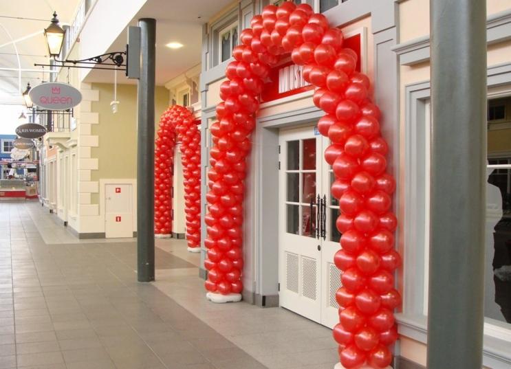 Czerwone balony uformowane w łuki dekorujące wejścia do lokali w pasażu handlowym.