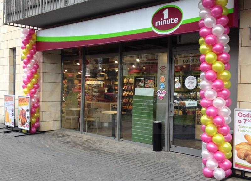 Wejście do sklepu '1 minute' ozdobione kolumnami z różowych, białych i żółtych balonów; w tle widoczne wnętrze sklepu z regałami i stolikami.
