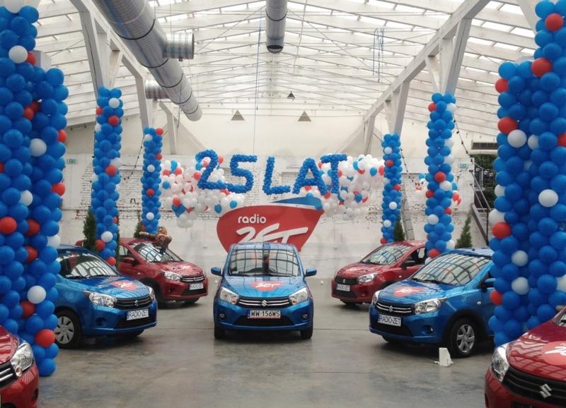 Uroczysta ekspozycja samochodów w hali, dekoracja balonowa w kolorach niebieskim, białym i czerwonym z napisem '25 LAT radio ZET', samochody z logo stacji radiowej.