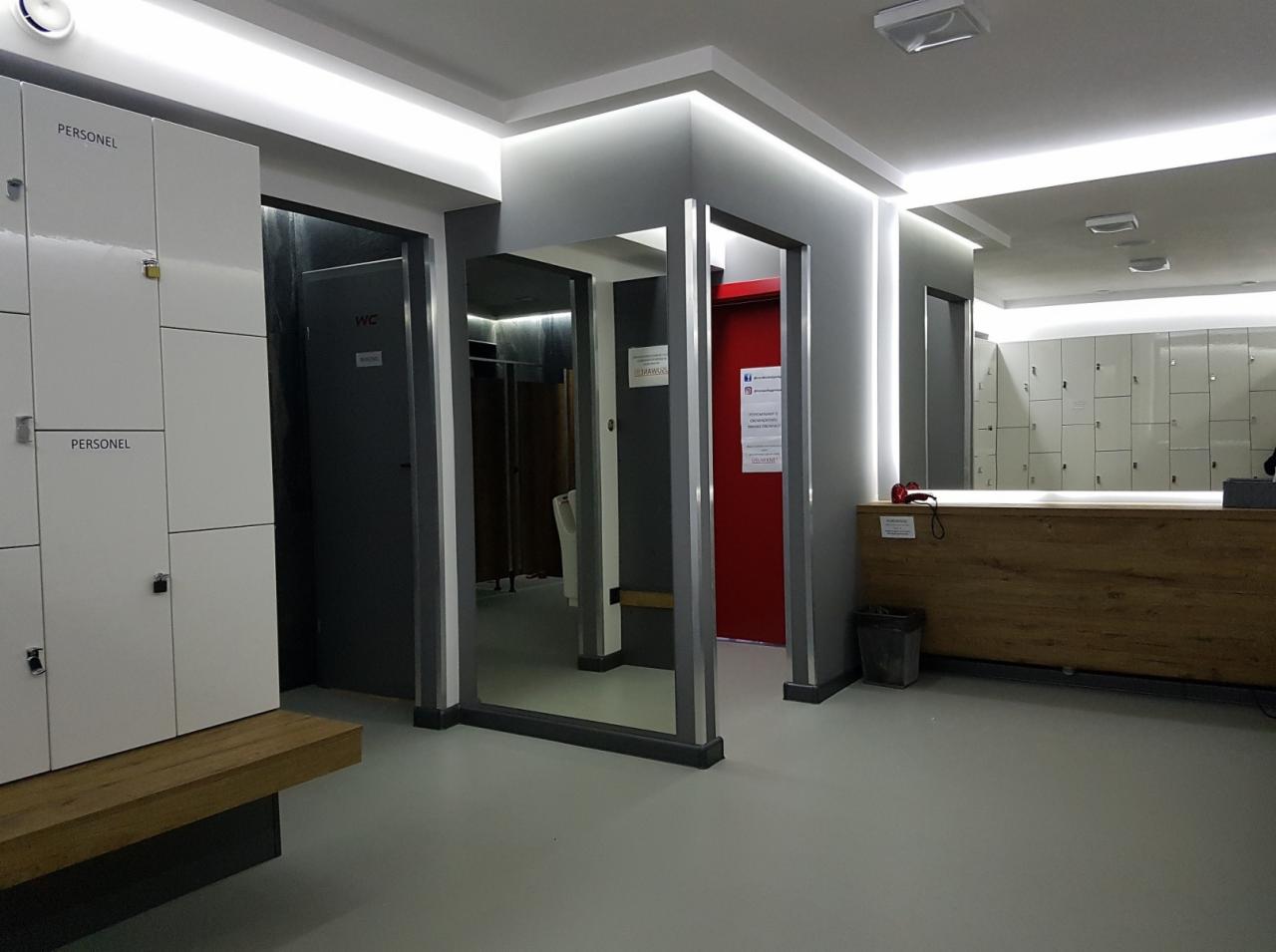 Nowoczesna szatnia pracownicza z białymi szafkami, drewnianą ławką i recepcją z drewnianym blatem, oświetleniem LED w suficie i szarymi ścianami.