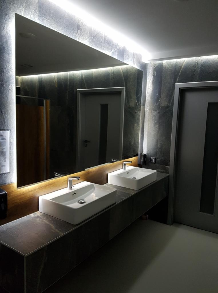 Nowoczesna łazienka publiczna z dwoma białymi umywalkami nablatowymi, kamiennym blatem i dużym lustrem z podświetleniem LED, szare płytki na ścianach i drzwi.