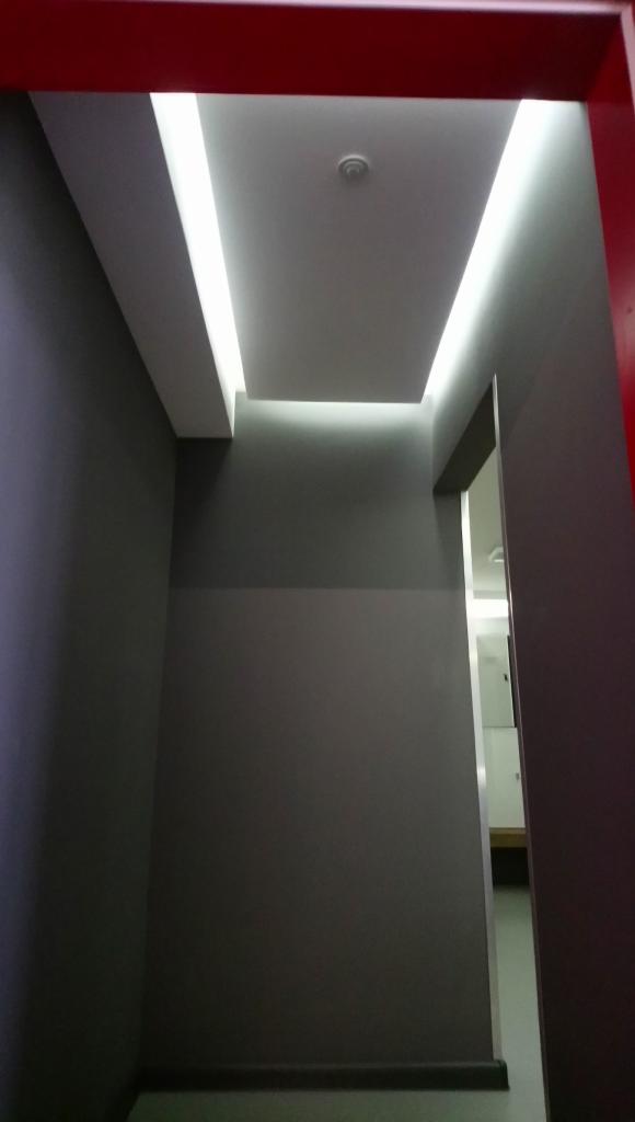 Korytarz z szarymi ścianami i podświetlanym sufitem podwieszanym z listwami LED, widok z dołu przez czerwone obramowanie drzwi.
