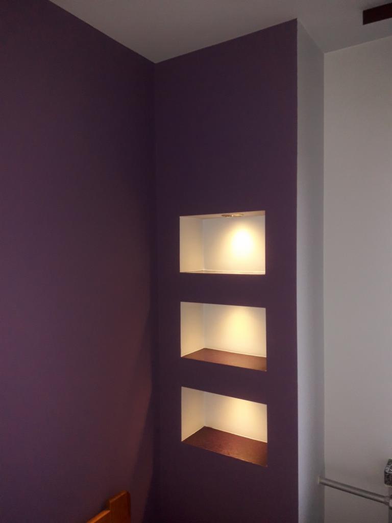 Fioletowa ściana z trzema podświetlanymi wnękami z drewnianymi półkami, widok z dołu, oświetlenie punktowe wewnątrz wnęk.