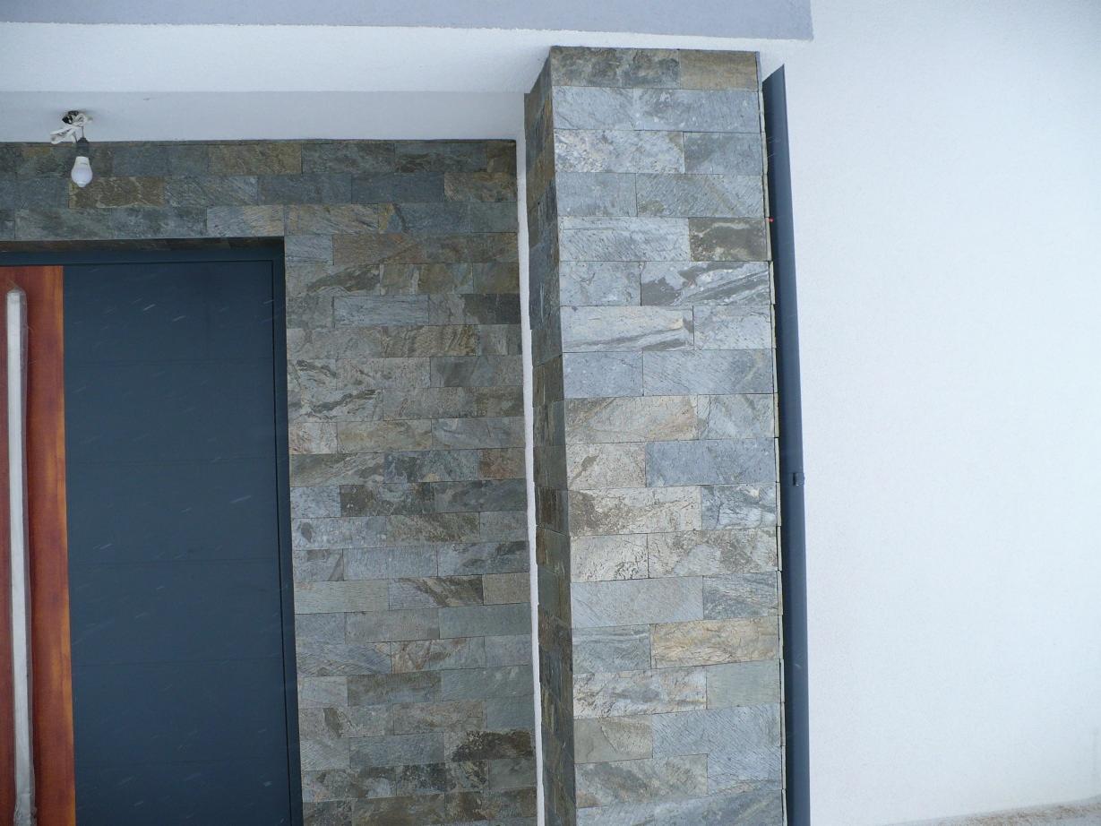 Ciemnoszare drzwi wejściowe z drewnianą ramą, otoczone dekoracyjną okładziną z kamienia naturalnego o nieregularnej fakturze i szaro-beżowej kolorystyce, z widoczną instalacją elektryczną pod sufitem.