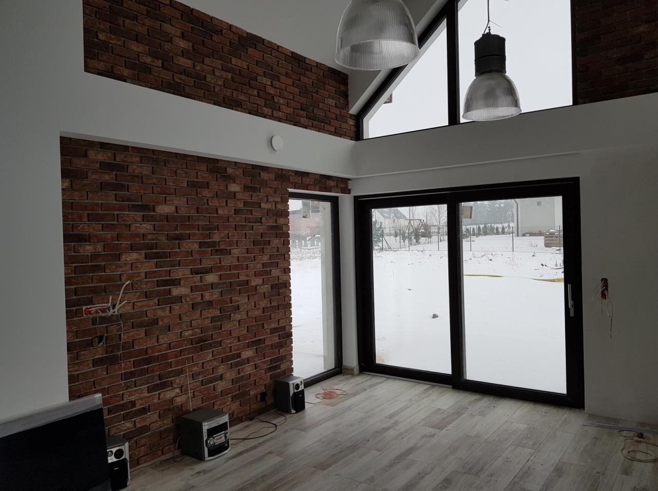 Surowe wnętrze w Wilkołazie z ceglaną ścianą, dużymi przesuwnymi oknami i drewnopodobną podłogą, oświetlone dwoma wiszącymi lampami w stylu industrialnym.
