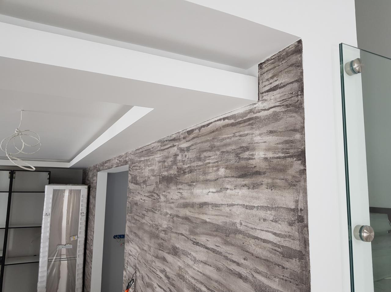 Ściana w łazience z dekoracyjnym malowaniem imitującym naturalny kamień, sufit podwieszany z oświetleniem LED, szklana ścianka kabiny prysznicowej z chromowanymi mocowaniami.