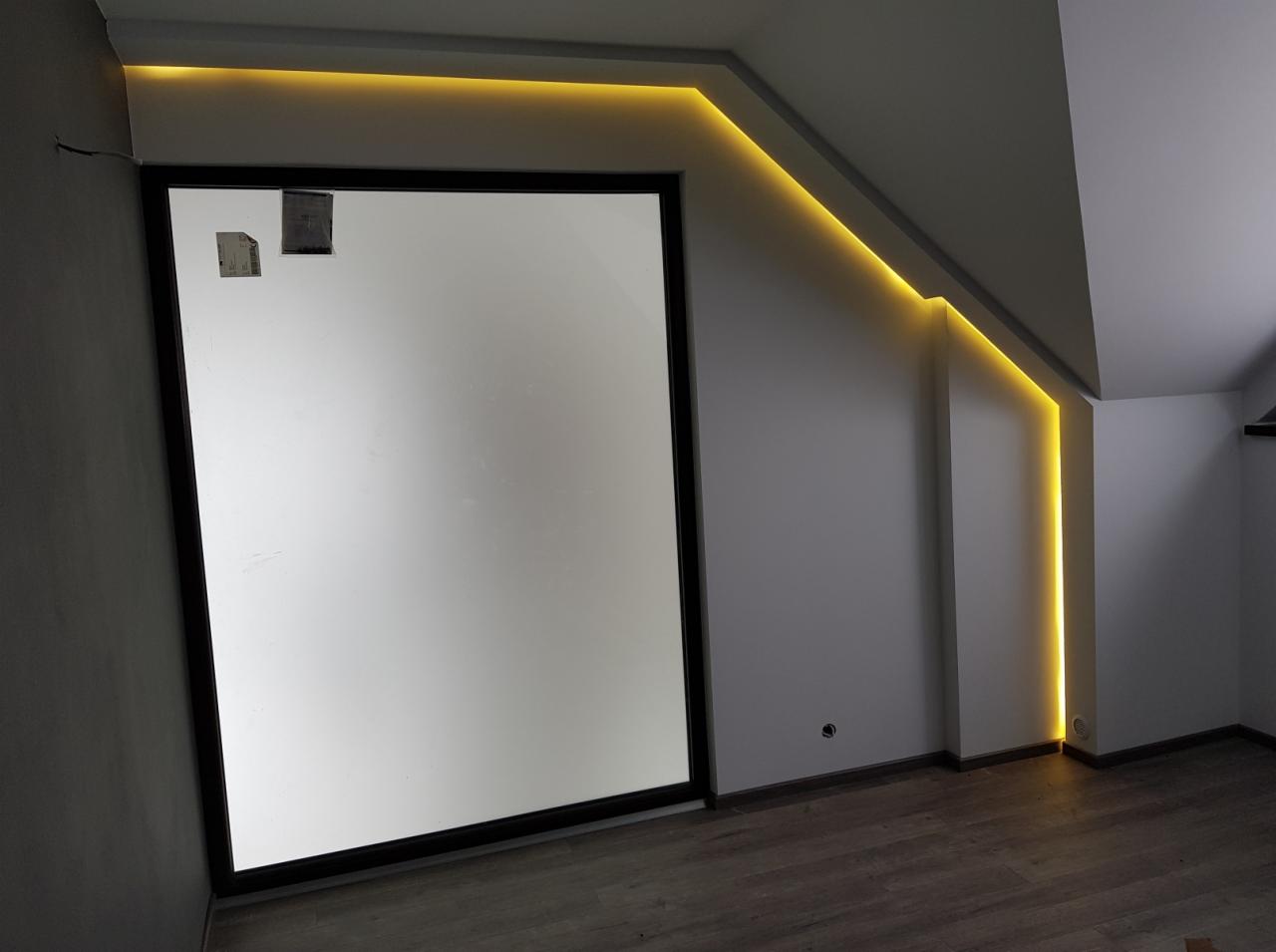 Pokój na poddaszu z oknem w czarnej ramie i dekoracyjnym oświetleniem LED wzdłuż krawędzi sufitu i ściany, widoczne ślady po taśmie malarskiej na szybie.
