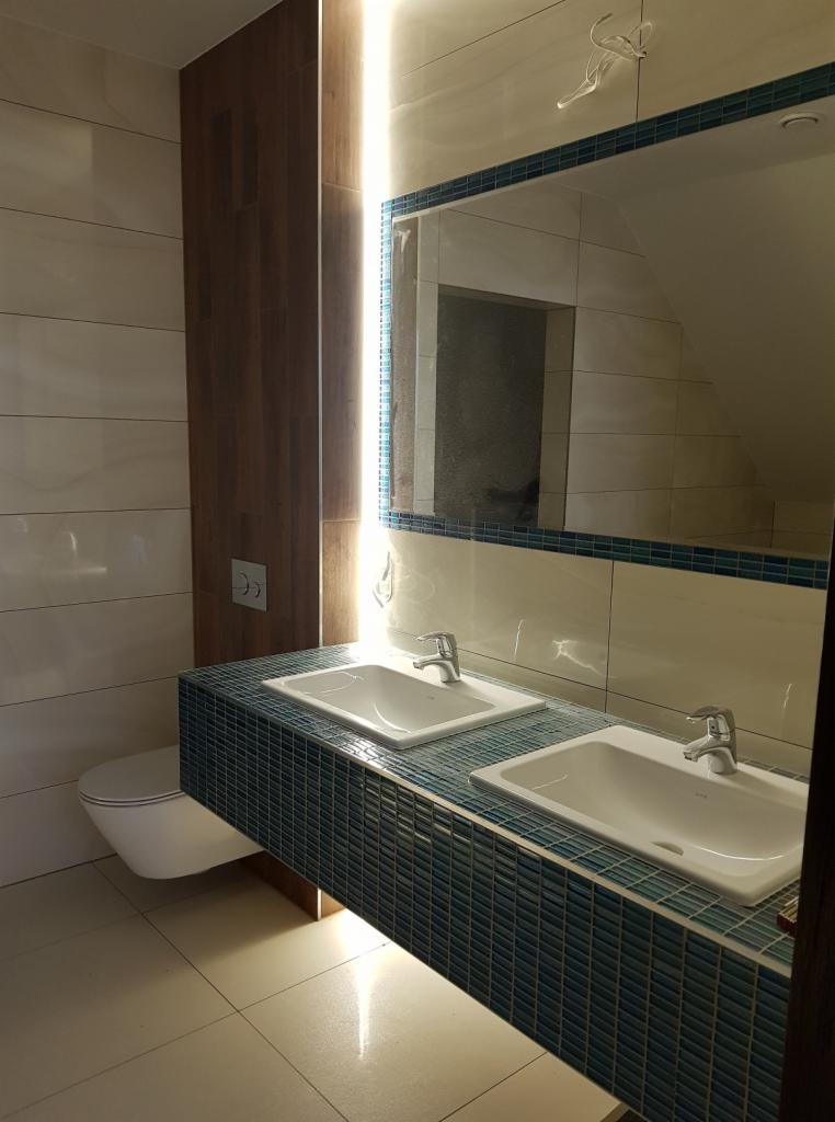 Nowoczesna łazienka z podwójną umywalką na blacie wykończonym mozaiką w odcieniach błękitu, lustro z niebieską obwódką i podświetleniem LED, oraz wisząca toaleta.