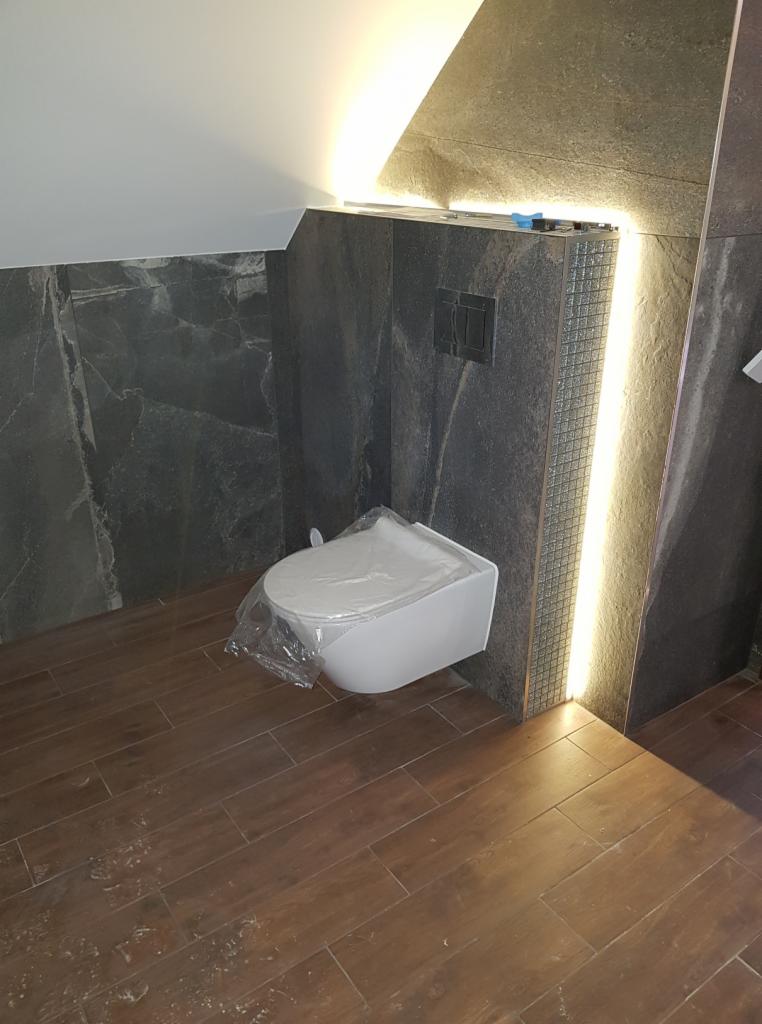 Nowa, podwieszana toaleta z przyciskiem spłukującym, zabezpieczona folią, na tle ściany z ciemnego kamienia z oświetleniem LED i podłogi imitującej drewno.